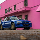 a blue vintage subaru 2.5rs coupe parked in front of a bright pink building on an overcast day. the subaru is built for rally racing, with oversized all-terrain tires and fender flares and scoops. a roll cage can be seen through the car's windows